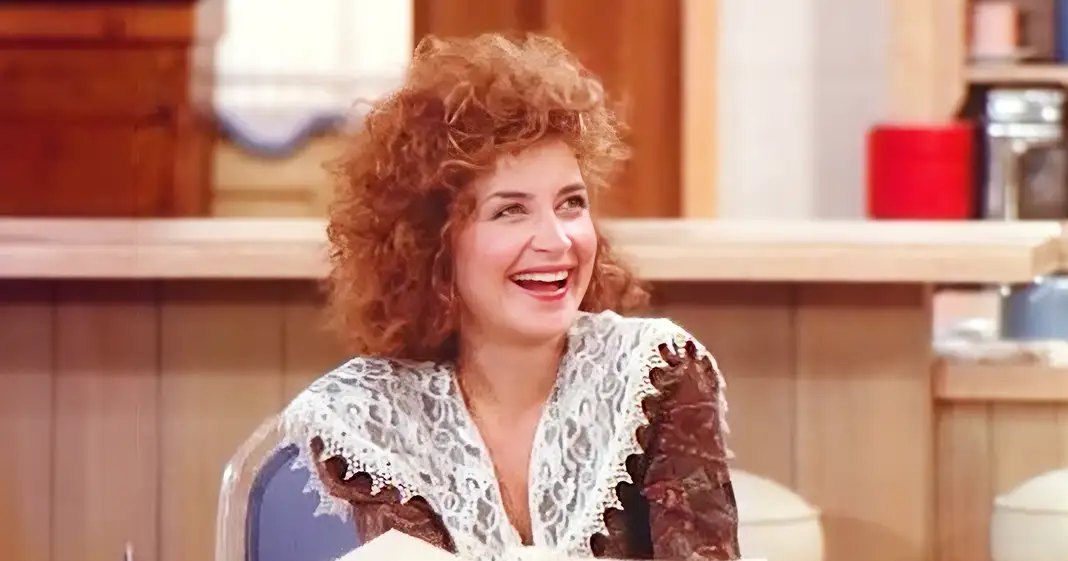 A middle-aged adult female wearing a white lace top with brown patterns and long sleeves, standing in an indoor kitchen or café setting with bright, natural lighting.