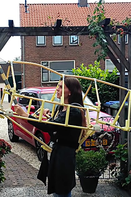 A young adult female holds a model airplane in an outdoor garden area with brick buildings, potted red flowers, and a decorative metal fence.