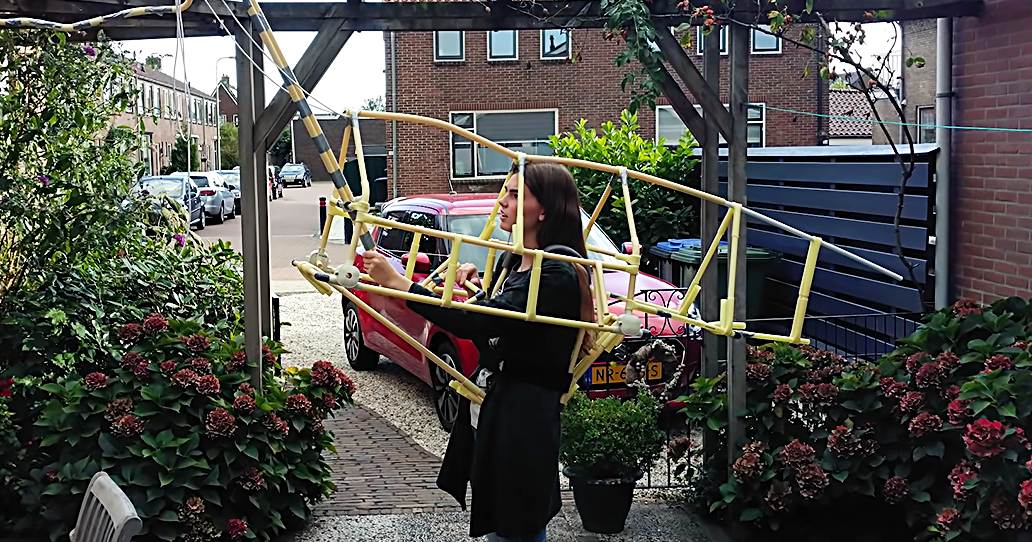 A young adult female holds a model airplane in an outdoor garden area with brick buildings, potted red flowers, and a decorative metal fence.