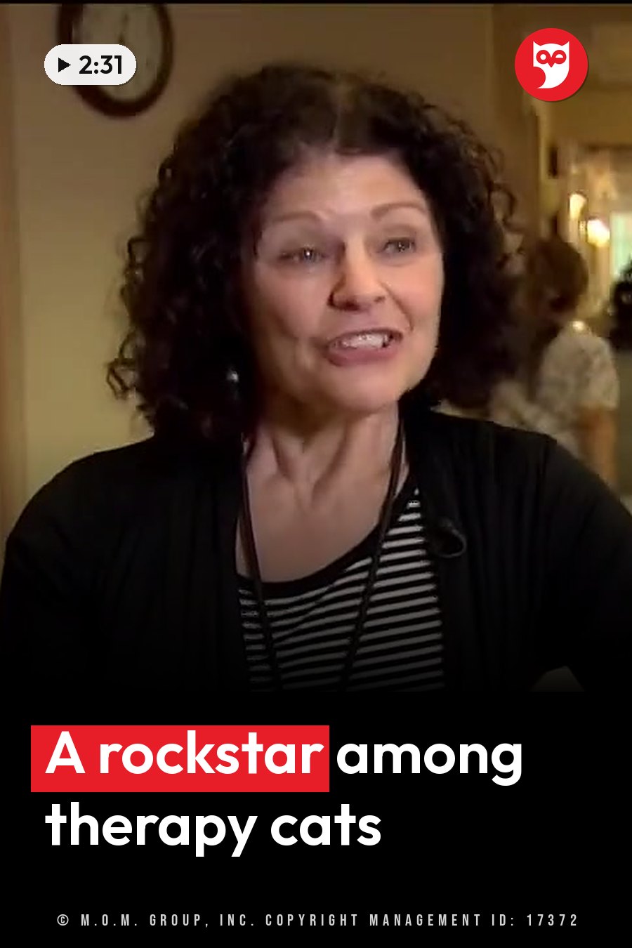 A woman with curly hair, in a striped shirt under a black cardigan, speaks indoors. A headline runs in white letters across the lower third.