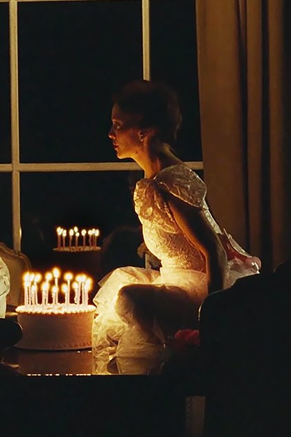 A young adult male in a striped sweater vest over a dark shirt and jeans stands next to an adult female wearing an elegant white gown with puffed sleeves, in a warmly lit, ornate dining room or living area.