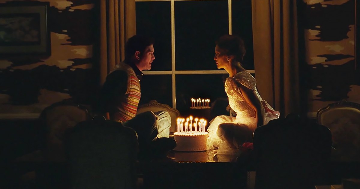 A young adult male in a striped sweater vest over a dark shirt and jeans stands next to an adult female wearing an elegant white gown with puffed sleeves, in a warmly lit, ornate dining room or living area.
