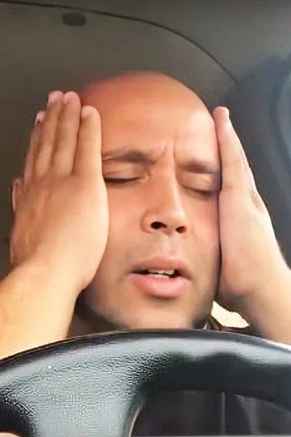 A middle-aged to older adult male wearing a dark-colored t-shirt with a simple design sits inside a car, showcasing part of the steering wheel and dashboard.