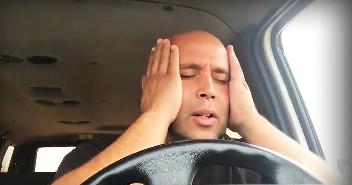 A middle-aged to older adult male wearing a dark-colored t-shirt with a simple design sits inside a car, showcasing part of the steering wheel and dashboard.