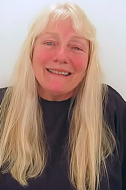 An older adult female in a relaxed, loose-fitting long-sleeved top or shirt with a round neckline against a plain white wall. Soft lighting suggests an indoor daytime setting.