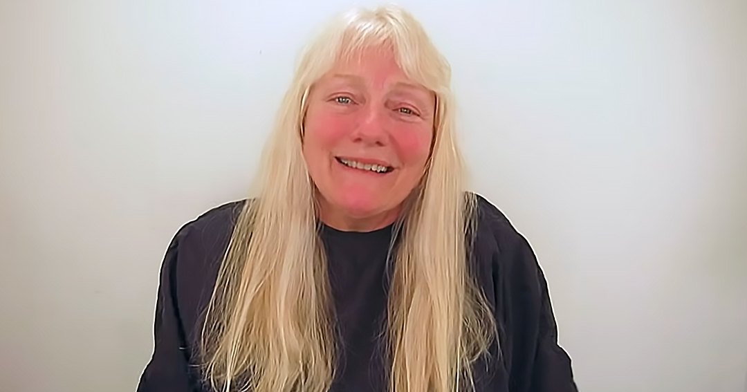 An older adult female in a relaxed, loose-fitting long-sleeved top or shirt with a round neckline against a plain white wall. Soft lighting suggests an indoor daytime setting.