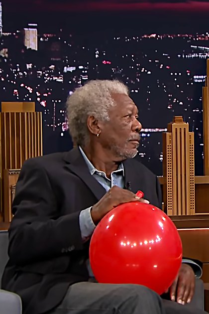 Two adults wearing suits, holding red balloons against a cityscape backdrop on 'The Tonight Show Starring Jimmy Fallon' set.