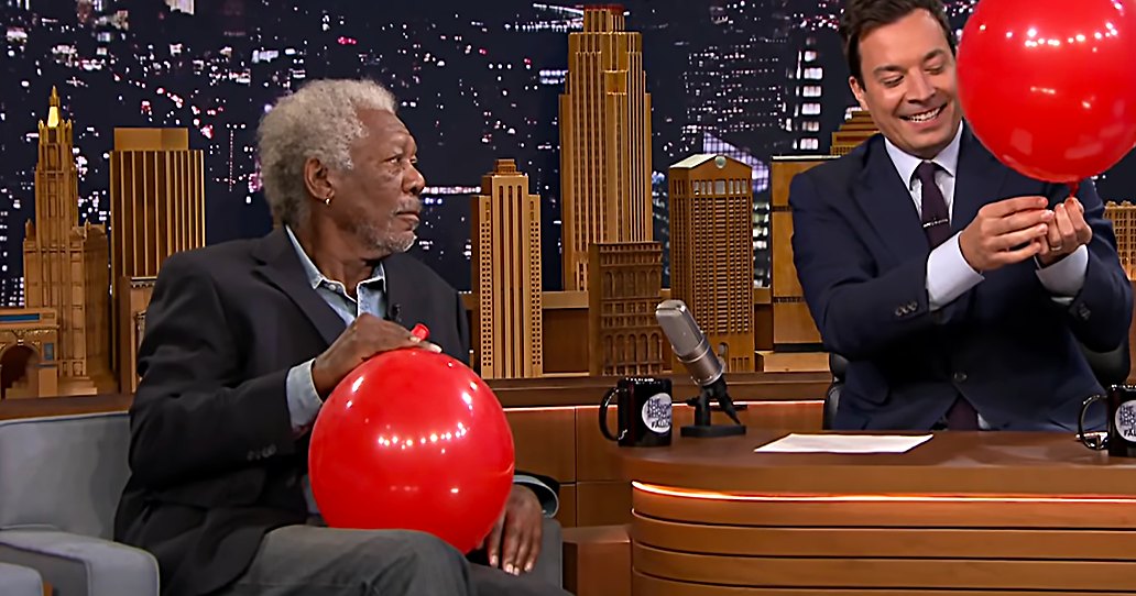 Two adults wearing suits, holding red balloons against a cityscape backdrop on 'The Tonight Show Starring Jimmy Fallon' set.