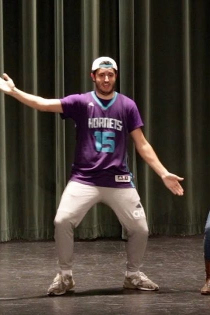 Young adult male performers in a school auditorium, wearing basketball jerseys and casual attire on stage with dark curtains behind them.
