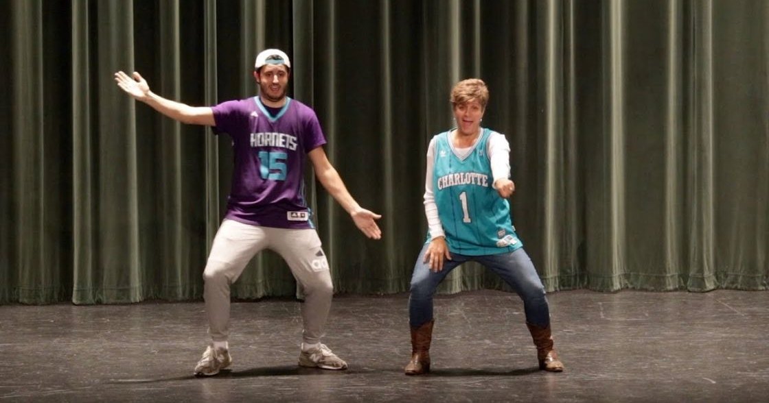 Young adult male performers in a school auditorium, wearing basketball jerseys and casual attire on stage with dark curtains behind them.