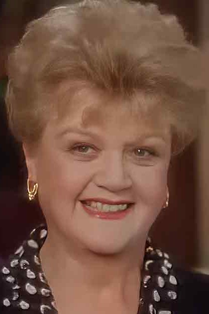 An older adult woman wearing a black top with white polka dots in an indoor social setting, surrounded by out-of-focus figures and greenery.