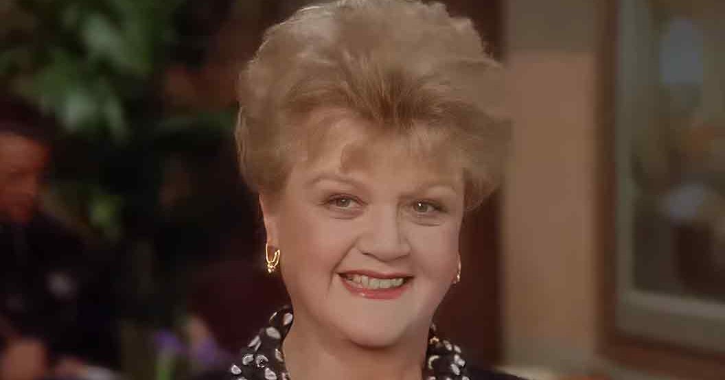 An older adult woman wearing a black top with white polka dots in an indoor social setting, surrounded by out-of-focus figures and greenery.