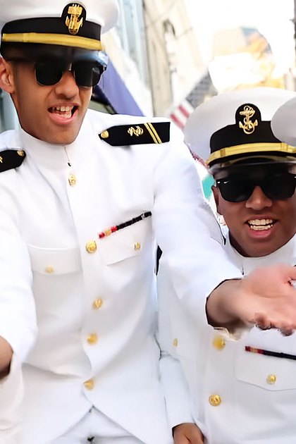 A group of young adults in white naval-style uniforms with gold buttons and black trims are gathered outdoors near a building with large windows, surrounded by greenery. The scene suggests an event or gathering during autumn daytime.