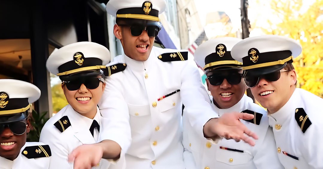 A group of young adults in white naval-style uniforms with gold buttons and black trims are gathered outdoors near a building with large windows, surrounded by greenery. The scene suggests an event or gathering during autumn daytime.