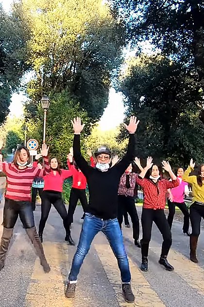 A group exercise class outdoors in a park-like setting with trees, paved path, and road signs indicating no entry; spring or summer foliage suggests daytime sunlight.