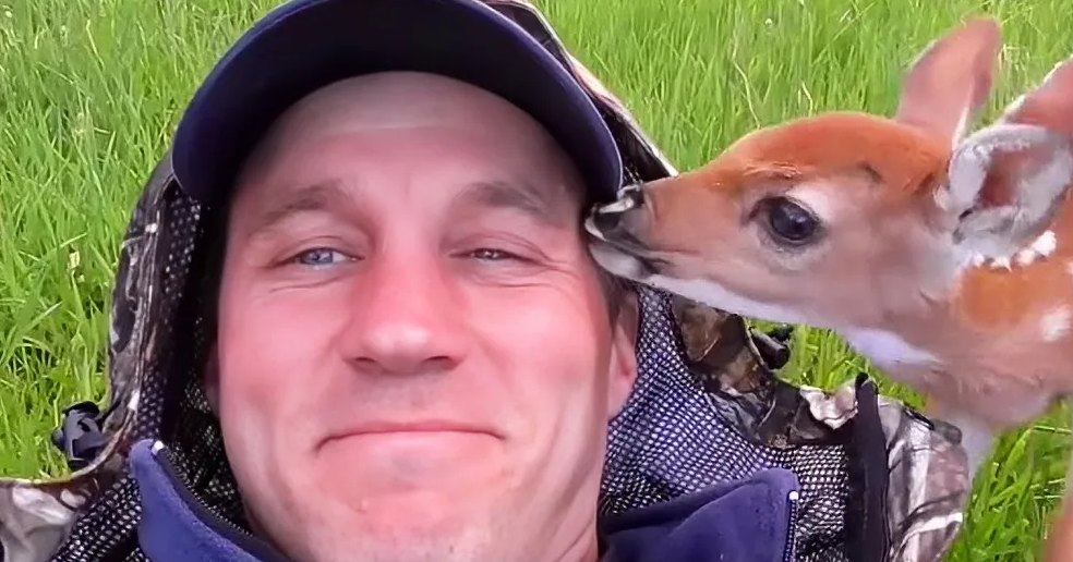 A man in a cap smiles as a fawn nuzzles his cheek in a grassy field.