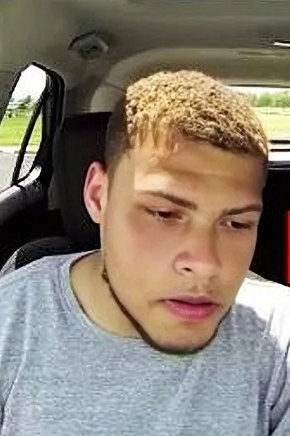 A young adult male in a car, wearing a gray t-shirt with cotton or cotton blend fabric, surrounded by an open area with grass and trees, suggesting spring or summer.