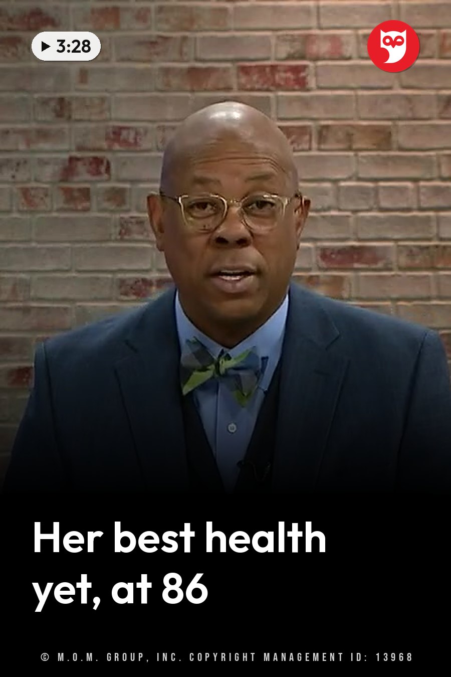 A bald man with glasses, a blue suit, a light shirt and a patterned bow tie speaks in front of a brick wall. A headline runs in white letters across the lower third.