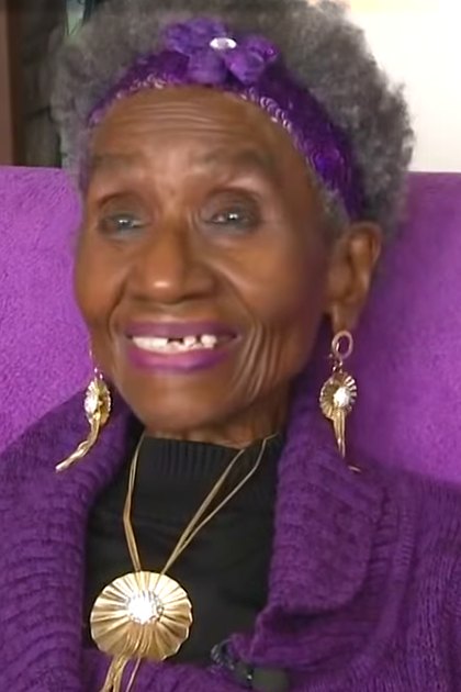 An older adult woman wearing a purple textured sweater and large hoop earrings sits indoors with a purple cushioned chair.