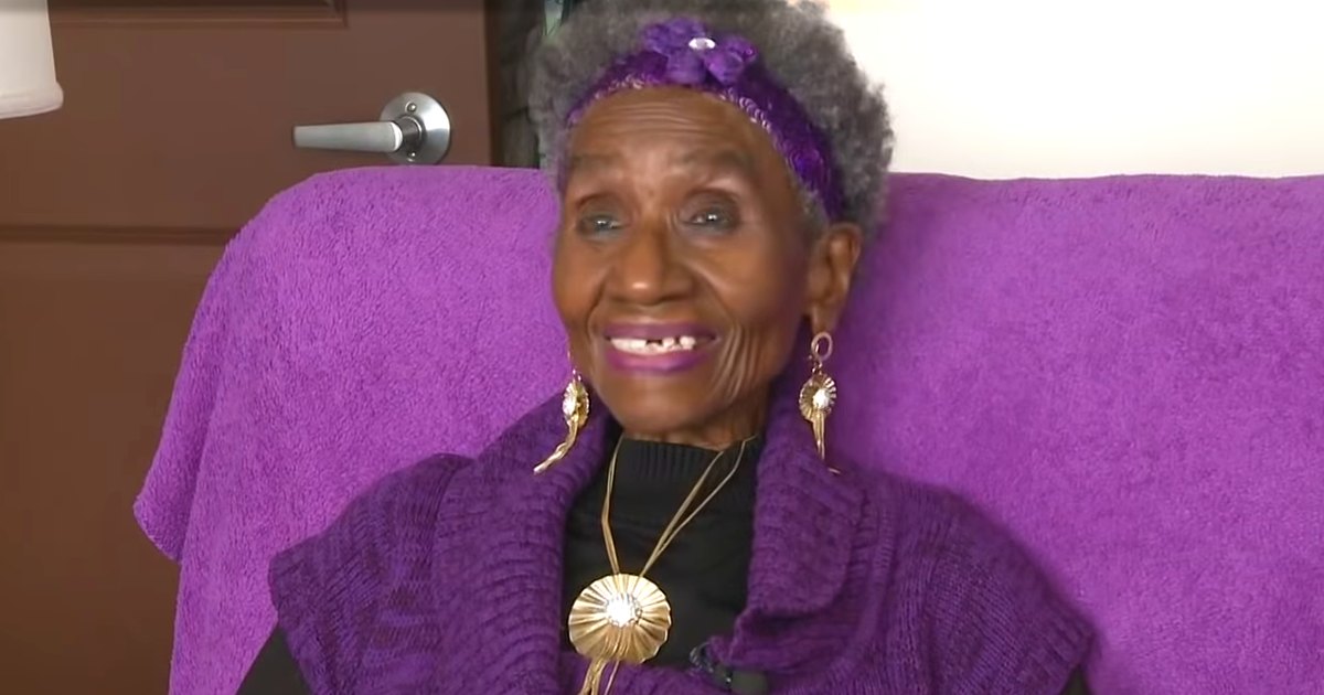 An older adult woman wearing a purple textured sweater and large hoop earrings sits indoors with a purple cushioned chair.