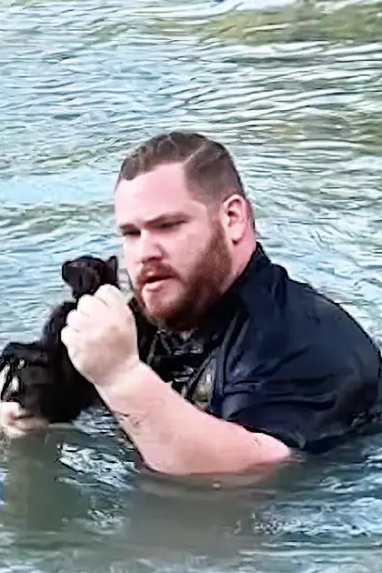 An adult male holds a small dark-furred dog near water, suggesting outdoor recreation in natural daylight.