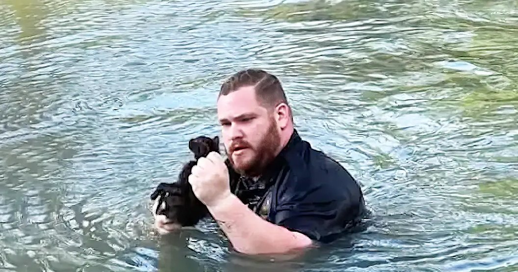 An adult male holds a small dark-furred dog near water, suggesting outdoor recreation in natural daylight.