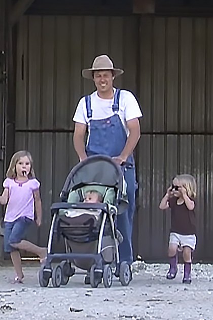 An adult male wearing overalls and a straw hat stands with children in front of a barn setting. The ground is sandy, scattered with small debris, suggesting farm life.