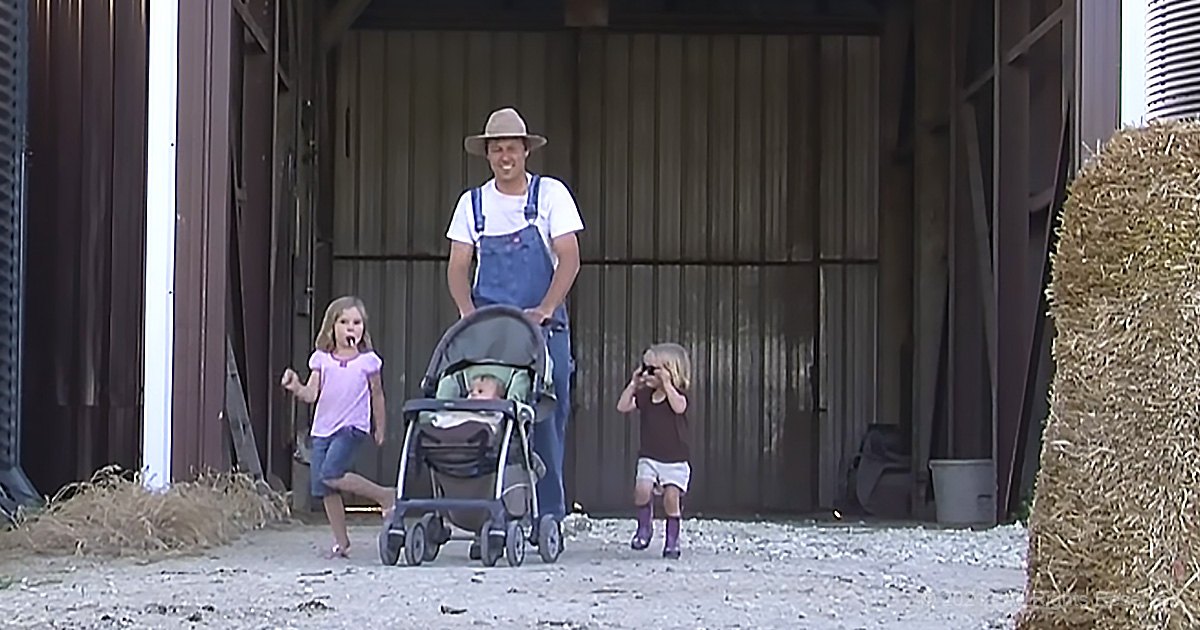 An adult male wearing overalls and a straw hat stands with children in front of a barn setting. The ground is sandy, scattered with small debris, suggesting farm life.