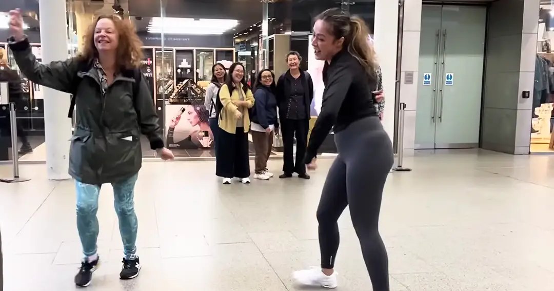 Two young adult females dancing in an indoor public space, possibly an airport terminal; casual attire and a line of onlookers standing nearby.