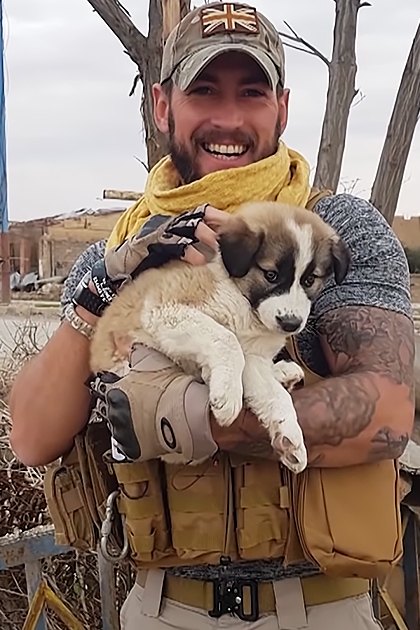An adult male holding a light brown and white puppy amidst damaged urban buildings, overcast late autumn or winter day with bare trees.
