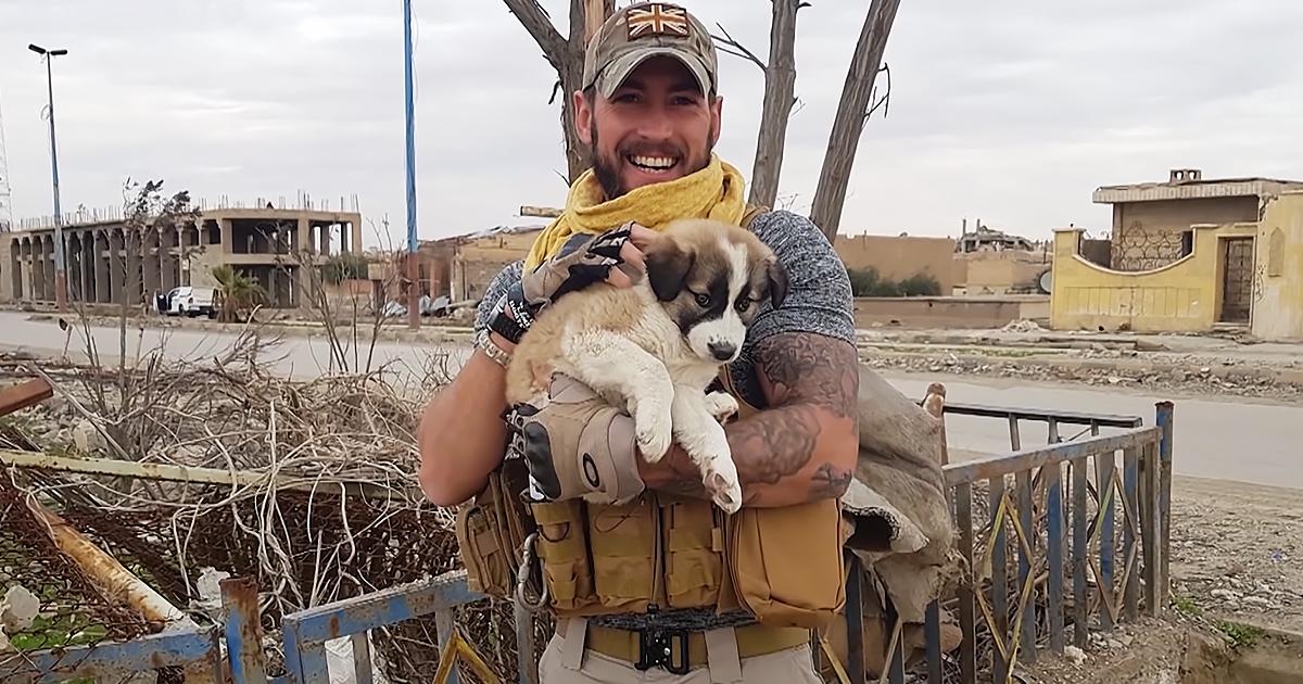 An adult male holding a light brown and white puppy amidst damaged urban buildings, overcast late autumn or winter day with bare trees.