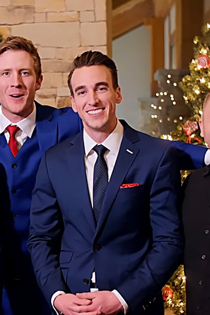 A group photo of young adult males in festive attire, including button-up shirts, suits with ties and pocket squares, gathered indoors against a backdrop decorated for the holidays. A guitar is held by one individual.