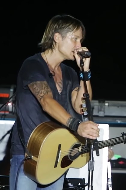 A young adult male singer wearing a dark t-shirt with a tattoo, holding a guitar, and a young adult female performer in a patterned dress with jewelry and bracelet, performing on stage at an indoor venue under artificial lighting.