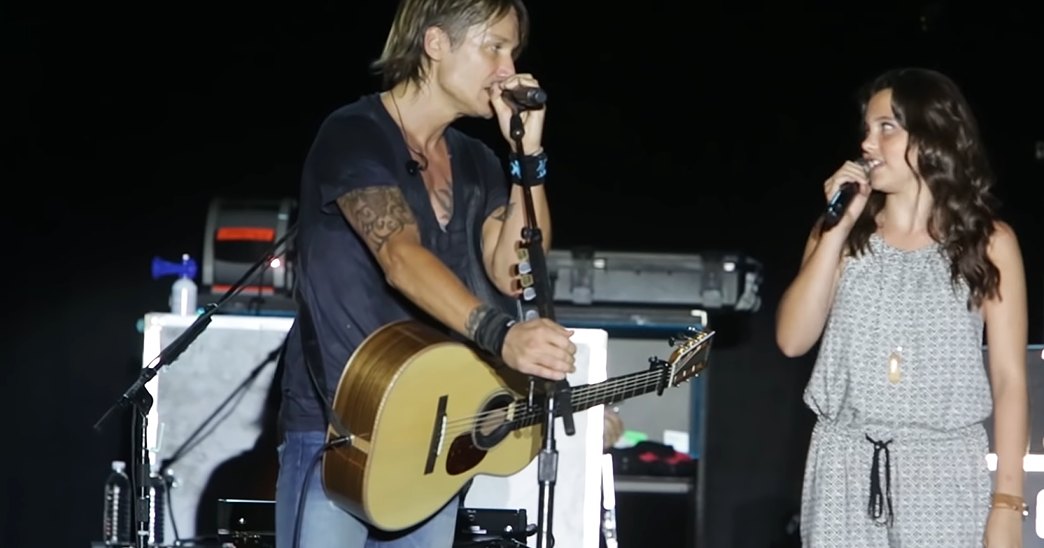 A young adult male singer wearing a dark t-shirt with a tattoo, holding a guitar, and a young adult female performer in a patterned dress with jewelry and bracelet, performing on stage at an indoor venue under artificial lighting.