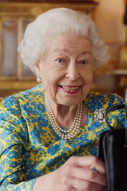 An older adult female wearing a blue and yellow floral dress with pearl jewelry, holding a black handbag, set against opulent grand palace decor.