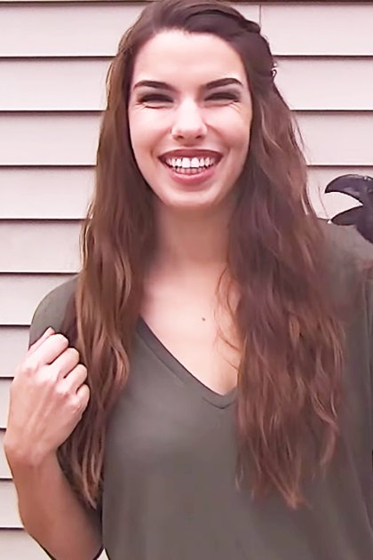 A young adult female wearing a dark green, relaxed-fit v-neck long-sleeved shirt outdoors in front of a house with light beige siding; a black-crow with white chest patch is present.