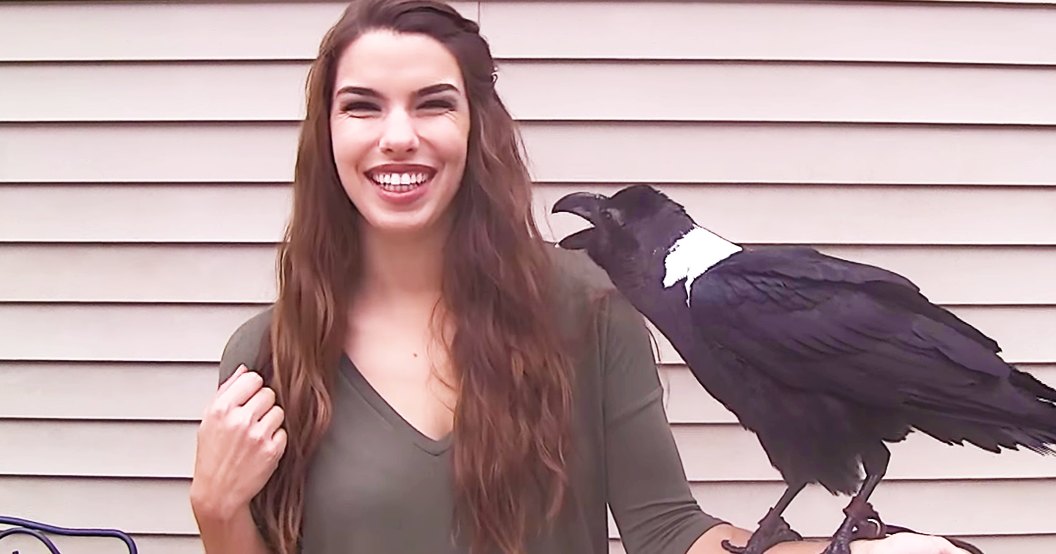 A young adult female wearing a dark green, relaxed-fit v-neck long-sleeved shirt outdoors in front of a house with light beige siding; a black-crow with white chest patch is present.