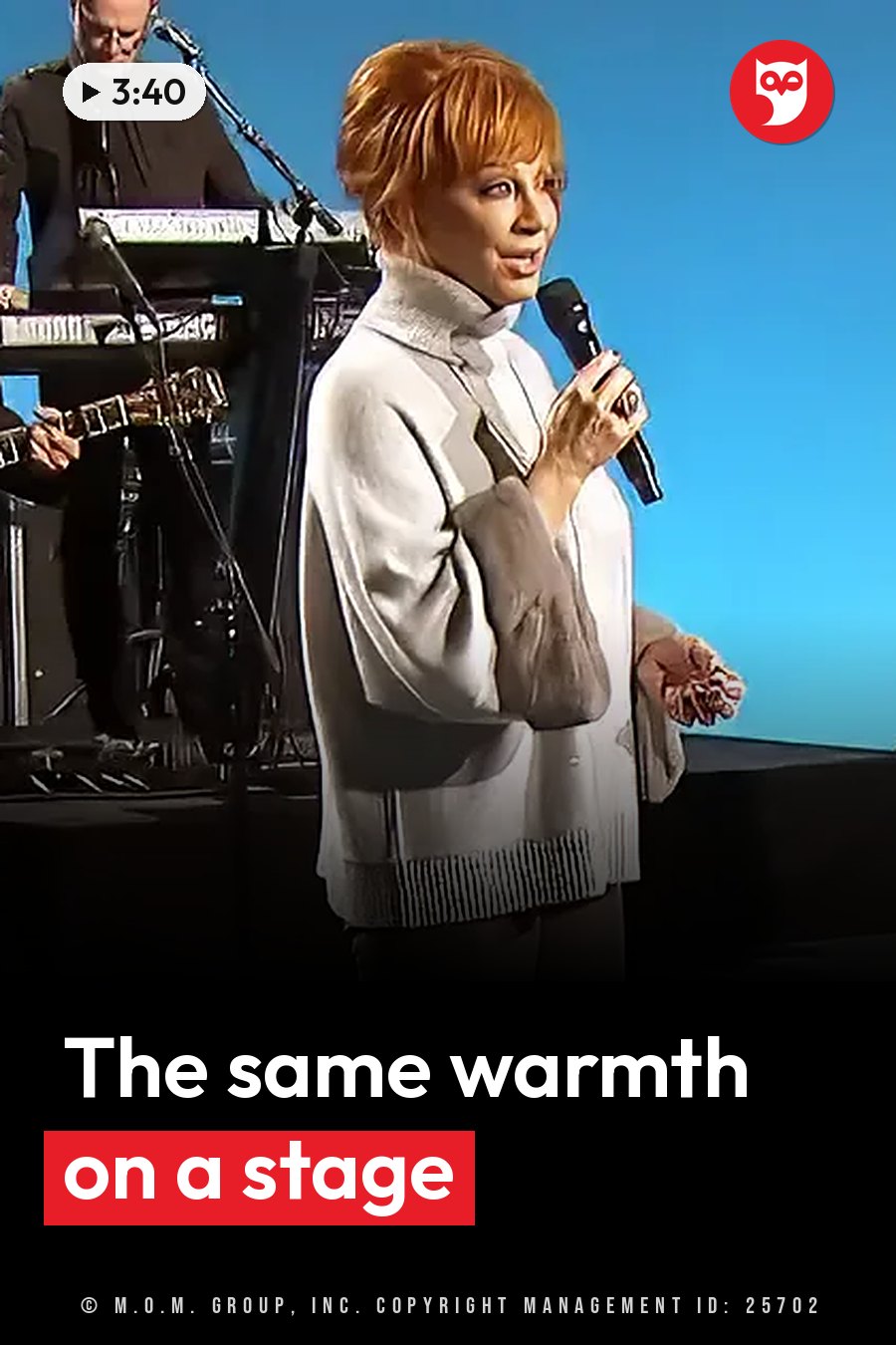A woman with short red hair holds a microphone while standing on a stage beside musical equipment. A headline runs in white letters across the lower third.