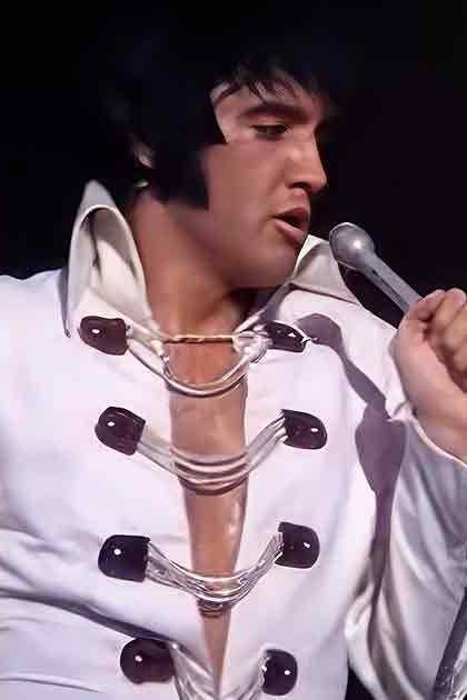 Adult male performer wearing a white tailored suit with large black buttons, holding a microphone during what appears to be a concert setting at night.