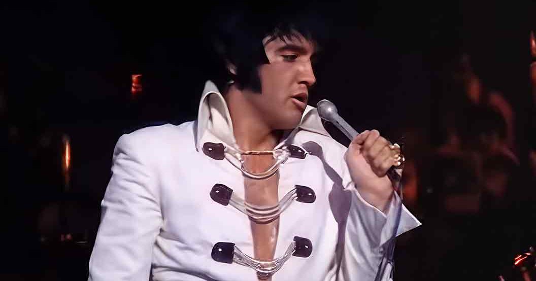 Adult male performer wearing a white tailored suit with large black buttons, holding a microphone during what appears to be a concert setting at night.