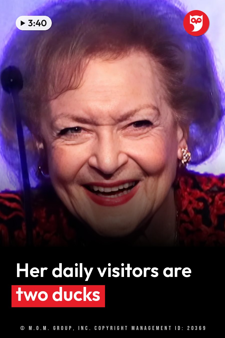 An older woman with styled hair, red lipstick and an ornate red outfit smiles broadly near a microphone. A headline runs in white letters across the lower third.