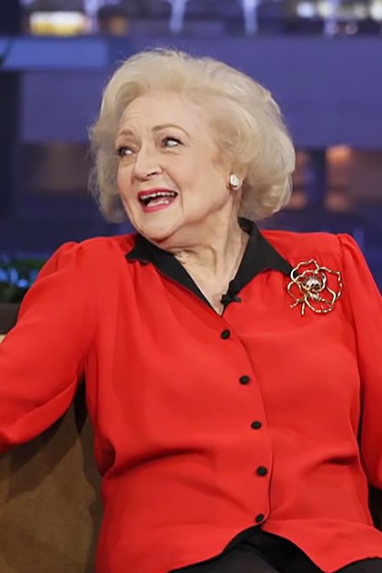 A young adult male guest and an older female talk show host wearing formal attire, seated on a brown couch with plants in the background.