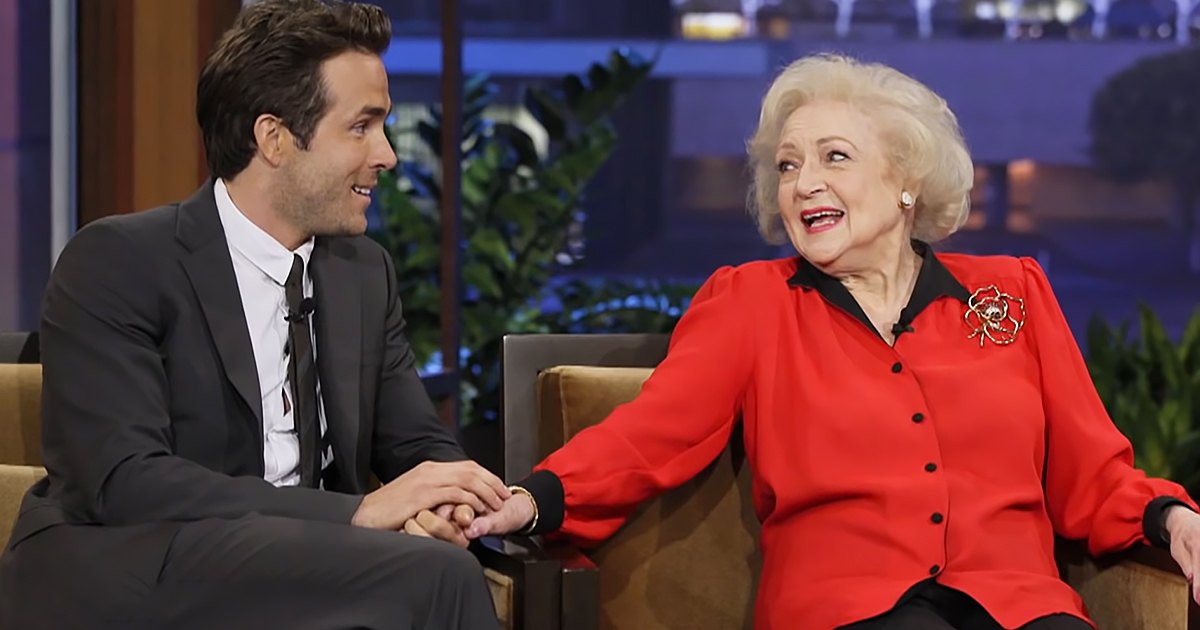 A young adult male guest and an older female talk show host wearing formal attire, seated on a brown couch with plants in the background.