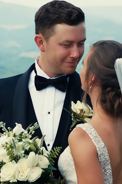 A young adult male singer in a black tuxedo and a young adult female wearing a white wedding dress with a long veil, holding a bouquet of white flowers against an overcast mountainous backdrop.