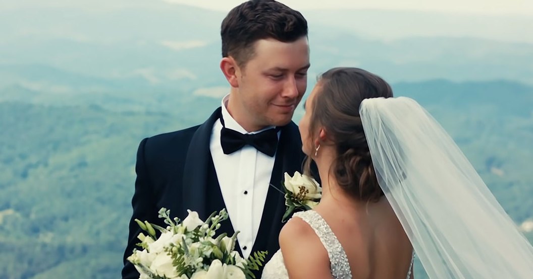 A young adult male singer in a black tuxedo and a young adult female wearing a white wedding dress with a long veil, holding a bouquet of white flowers against an overcast mountainous backdrop.