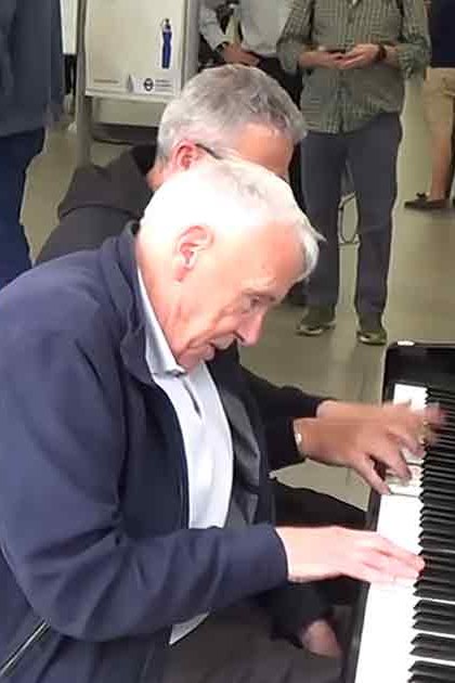 A man playing a piano on a train station platform with other people waiting in the background.