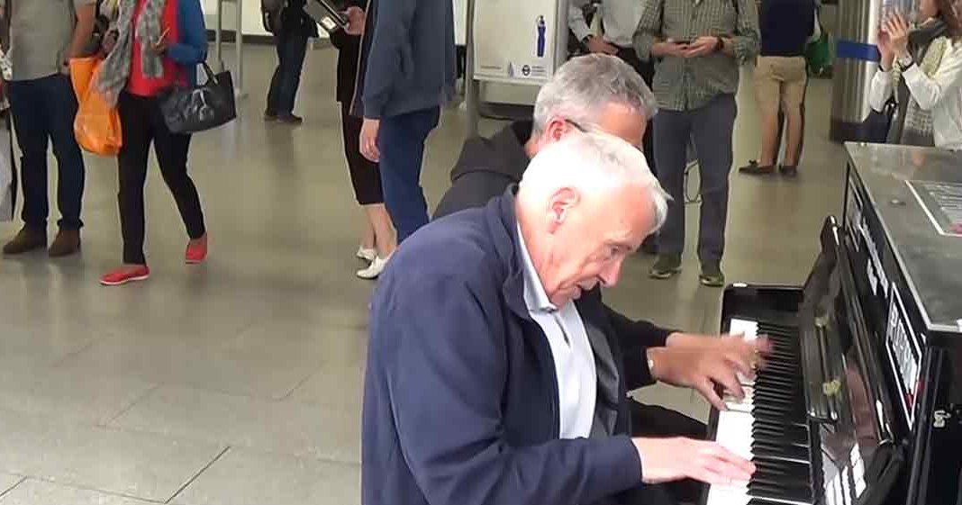 A man playing a piano on a train station platform with other people waiting in the background.