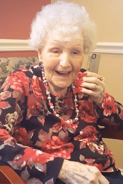 An older adult woman in a floral blouse with red flowers, sitting on an ornate chair indoors at a nursing home. A younger adult staff member is present nearby.