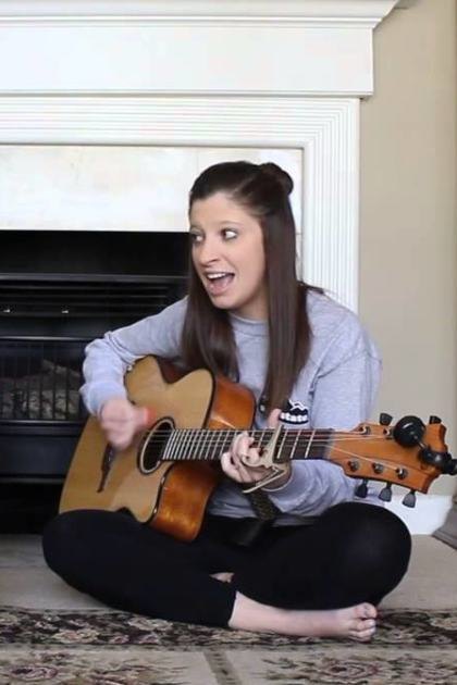 Young adult male singer in maroon sweatshirt, young adult female barefoot in gray shirt and black leggings, cozy living room setting with white fireplace and wooden cabinets.