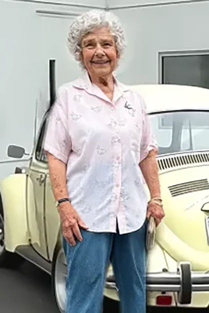 An older adult female wearing a light pink button-up shirt with small floral patterns stands next to a vintage Volkswagen Beetle at a service center, holding hands with an adult male in a light gray hoodie and black cap.